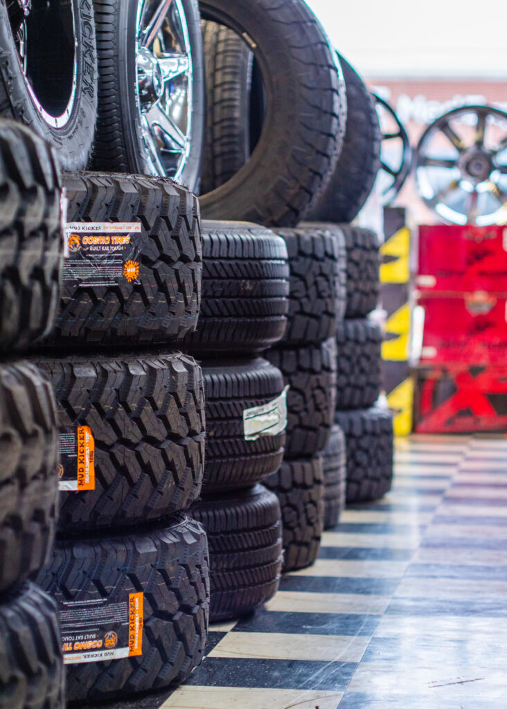 Four stacks of different types of tires are lined up in a shop. The shop has black-and-white checkered tiles.