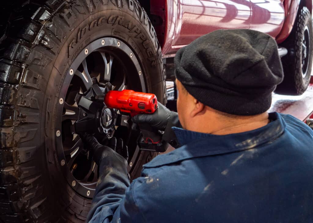 A Robert Jr's Tire & Auto employee in dark pants, a blue jacket, and a black beanie hat is using a lug drill on a lifted red truck.